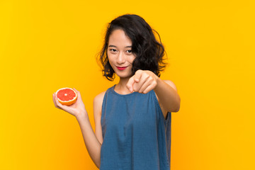 Young asian girl holding a grapefruit over isolated orange background points finger at you with a confident expression