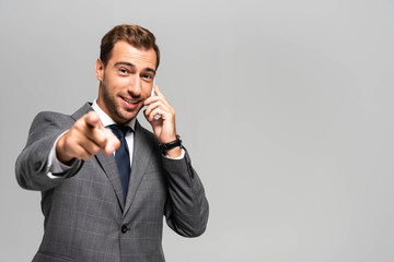 handsome and smiling businessman in suit talking on smartphone and pointing with finger isolated on grey