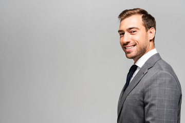 smiling and handsome businessman in suit looking at camera isolated on grey