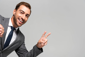 handsome and smiling businessman in suit showing peace sign isolated on grey