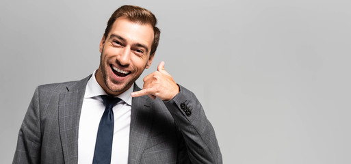 panoramic shot of smiling businessman in suit showing call me sign isolated on grey