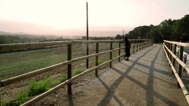 Girl with long hair in black kaftan dress and black hat leans on a fence on a rural road and looks at a beautiful view. Sunny day at countryside. Elegant young woman in black hat. Fashion and beauty.