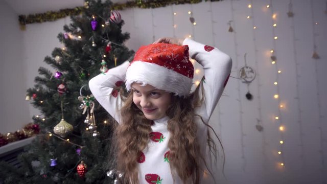 little girl in santa claus hat. the child adjusts the cap with the help of a pompom. curls hair with a finger and fools around.