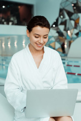 attractive and smiling woman in white bathrobe using laptop in spa