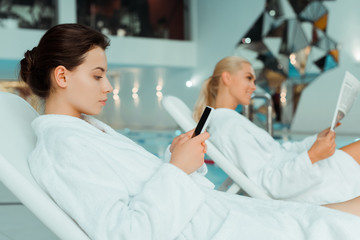 selective focus of attractive woman using smartphone and friend reading newspaper on background in spa
