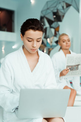selective focus of attractive woman using laptop and friend reading newspaper on background in spa