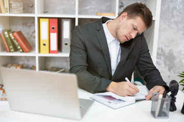 Naklejka premium busy man manager with laptop sitting at his working place in office taking notes in planner and having phone conversation looking confident. multitasking, work concept