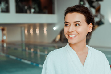 attractive and smiling woman in white bathrobe looking away in spa