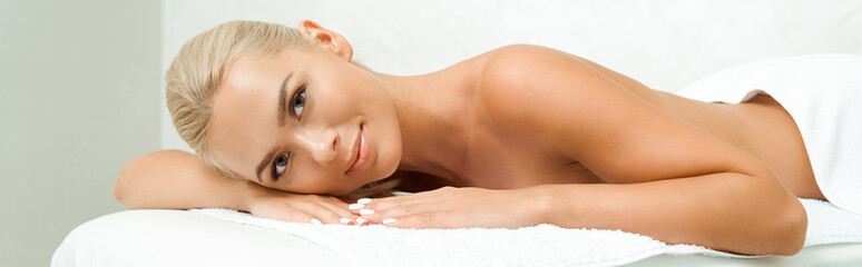 panoramic shot of attractive and smiling woman looking at camera and lying on massage table in spa