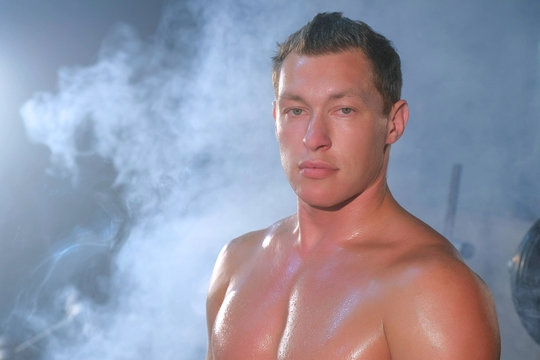 Portrait of professional muscular athletic man bodybuilder is sitting on bench in gym and looking at camera. Perfect body with naked torso. White smoke in gym in dark. Sport motivation concept.