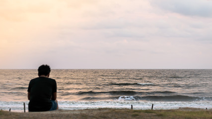 Blur image of a solitary man sitting alone at the beach in sunset time. Rear View of a lonely thinking person in wilderness area. Rest, relax, leisure activity background concept.
