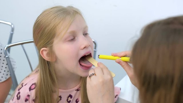 Medicine for children and healthcare concept. Pediatrician looking child girl throat using spatula and lantern, portrait of child. Preventive examination in clinic. Diagnostics tonsillitis or angina.
