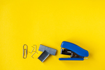 Blue stapler with metal staples and paper clip on yellow background. Top view. Copy, empty space for text. Minimalistic concept - obrazy, fototapety, plakaty