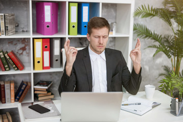 Naklejka premium young handsome man manager sitting with his fingers crossed hoping for successful negotiations at his working place in office. multitasking, work concept