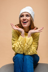 beautiful positive woman in yellow sweater and hat sitting on armchair, isolated on beige