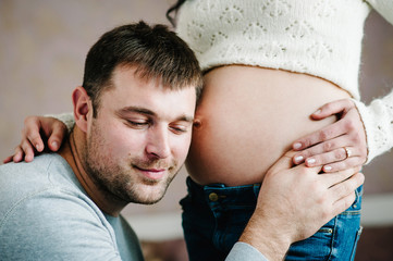 Handsome man is listening to his beautiful pregnant wife's tummy and smiling. Woman and loving man hugging tummy at home. Loving Couple. Parenthood concept. Baby Shower.