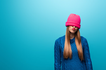 attractive offended woman in knitted sweater with pink hat on eyes, isolated on blue