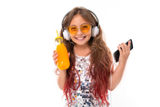 Little pretty caucasian girl listen to music with big earphones and drinks orange juice, picture isolated on white background