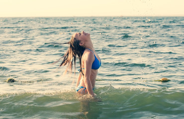 Young girl is standing in the sea and throwing her long wet hair back, water splashing in the air, with blue sky and copy space