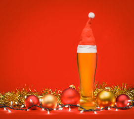 Tall glass of beer with red santa hat christmas baubles tinsel and lights on red background