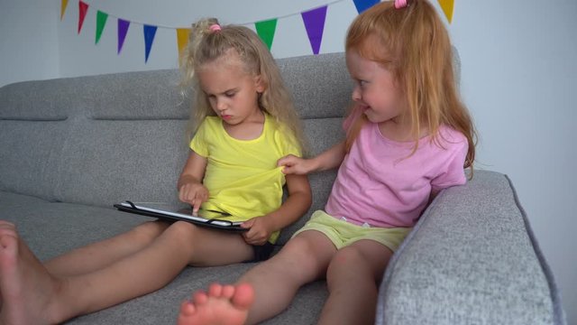 Addictive children girls fighting for tablet computer. Best friends on sofa