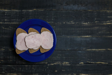 Obraz na płótnie Canvas Sandwich of dark bread and sausage slices on blue plate standing on black wooden background. Tasty unhealthy food. Diet, nutrition, eating habits concept. Place for text
