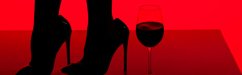 cropped view of black silhouette of sensual girl in heels with glass of wine isolated on red, panoramic shot