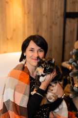 A beautiful and happy woman of 50 years old is sitting with a cat in her arms and under a plaid on a sofa. Christmas 2020.