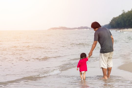 Portrait image of Asian Father with his daughter are​ hand​ in​ hand​ and​ walking​ on the beach at the sea. Summer season. Father's day and family concept.