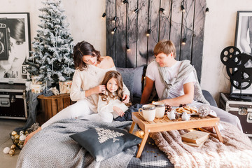Happy loving family mother, father and child on Christmas sit on the bed with a drink. New year 2020. Parents looking at daughter. Parents play with their daughter. Mom does a hairdress to her girly.