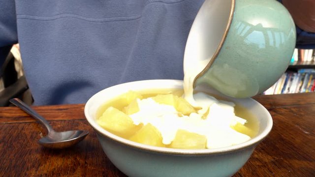 Closeup circular dolly shot of a man sitting at a wooden table, pouring cream from a jug onto a bowl of pineapple pieces.