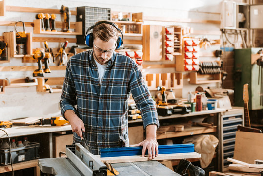 handsome woodworker in protective headphones holding plank near circular saw