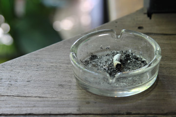 Cigarette butt with ash in glass ashtray on wooden table.