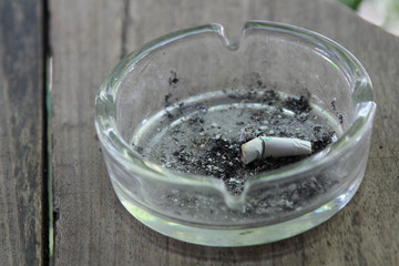 Cigarette butt with ash in glass ashtray on wooden table.