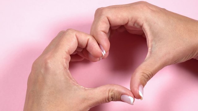 Close up view of two female caucasian hands isolated on pink pastel background. Young woman forming shape of heart with her fingers. Real time full hd video footage. Point of view shot.