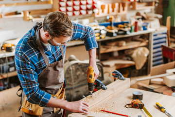 woodworker in goggles and apron holding hammer drill near wooden planks