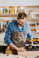 selective focus of carpenter in safety glasses and apron holding hammer drill near wooden planks