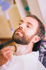 Close-up view of a young man with a beard in the barbershop. The barber's hand applies shaving foam...