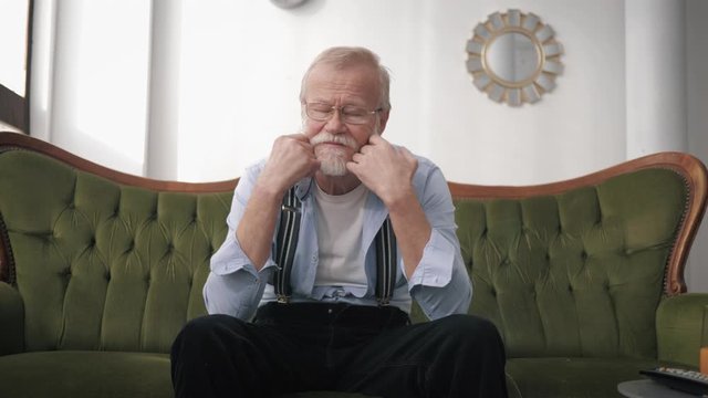 Depression, an old pensioner with a beard and glasses for vision suffers and is sad because of life difficulties sitting in a room on the couch