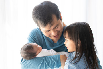 Fototapeta premium Asian young father is carrying his newborn baby boy and talking with his lovely daughter about her brother, concept of love and relation in family and bonding of the parent to their child, 
