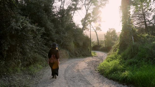 Girl with long hair in black kaftan dress and black hat walking along a rural road. Sunny day at countryside. Elegant young woman in black hat. Fashion and beauty.