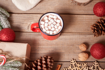 Red mug of Christmas cocoa with marshmallows, gift, cone, candy cane and Christmas decoration on the wooden table