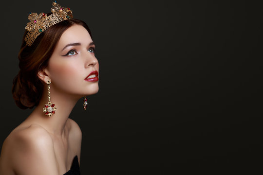 Beautiful girl with red lipstick in golden crown and earrings on dark background