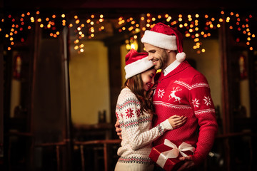 Happy couple in warm sweaters and santa hats embracing on street