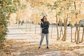 Fototapeta premium Stylish gorgeous young female photographer in a blue raincoat, jeans, black snickers, and sunglasses holds the camera in bent hand and waits for the model in the sunny forest in the afternoon