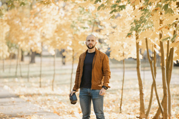 Bald brutal photographer with a beard in a suede leather jacket, blue shirt and jeans holds the camera and looks for a model in the park in the afternoon