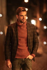 handsome, confident man in autumn outfit looking away and holding hand in pocket while standing on evening street