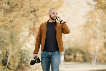 Bald stylish photographer with a beard in a suede leather jacket, blue shirt, jeans with analog wristwatch holds the camera and calls somebody by his smartphone in the park in the afternoon