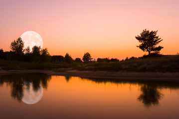 small quiet lake, tree silhouette and huge rising moon reflected in a water
