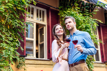 Fototapeta premium happy couple holding glasses of red wine while standing near house covered with green ivy and looking away